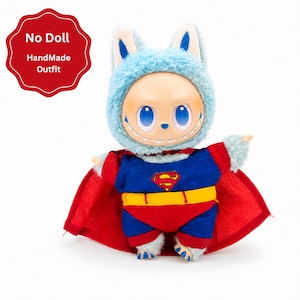 Puede incluir: Un disfraz hecho a mano para mu&ntilde;eca, con una capucha azul y peluda con orejas de conejo. La mu&ntilde;eca viste un disfraz de Superman con capa roja, body azul y rojo y cintur&oacute;n amarillo. El texto "No Doll Handmade Outfit" est&aacute; en un c&iacute;rculo rojo.