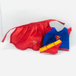 Puede incluir: Un conjunto de capa y pantal&oacute;n corto rojos con el logo de Superman, combinado con una camiseta azul y roja con el mismo emblema. Un cintur&oacute;n amarillo completa el disfraz de superh&eacute;roe. El conjunto es para un ni&ntilde;o.