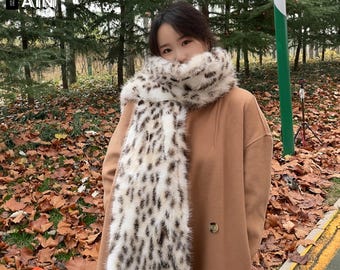May include: A cream, brown, and black spotted faux fur scarf. The scarf is worn over a tan coat. The image includes the text "TREND AIN" in the upper left corner. The background features autumn leaves and trees.