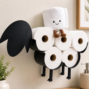 May include: A whimsical black metal toilet paper holder shaped like a rooster, holding six rolls of white toilet paper. A plush toilet paper roll with a smiling face and brown legs sits on top. The holder is mounted on a beige wall.