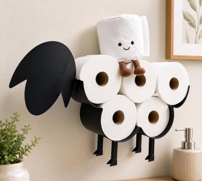 May include: A whimsical black metal toilet paper holder shaped like a rooster, holding six rolls of white toilet paper. A plush toilet paper roll with a smiling face and brown legs sits on top. The holder is mounted on a beige wall.