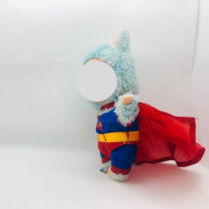Puede incluir: Un peluche disfrazado de superh&eacute;roe, con una cabeza azul claro y una capa roja. El disfraz incluye un traje azul y rojo con un cintur&oacute;n amarillo y un logotipo de Superman.