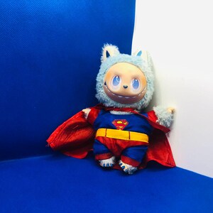 Puede incluir: Un juguete de peluche disfrazado de Superman, con un traje azul y rojo, un cintur&oacute;n amarillo y el logotipo de Superman. El juguete tiene una cabeza peluda azul claro con una cara sonriente y una capa roja. El fondo es una esquina azul y blanca.