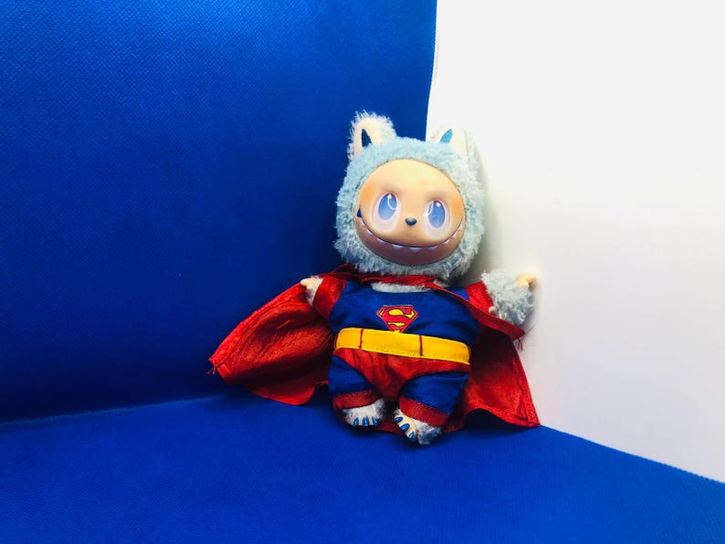 Puede incluir: Un juguete de peluche disfrazado de Superman, con un traje azul y rojo, un cintur&oacute;n amarillo y el logotipo de Superman. El juguete tiene una cabeza peluda azul claro con una cara sonriente y una capa roja. El fondo es una esquina azul y blanca.