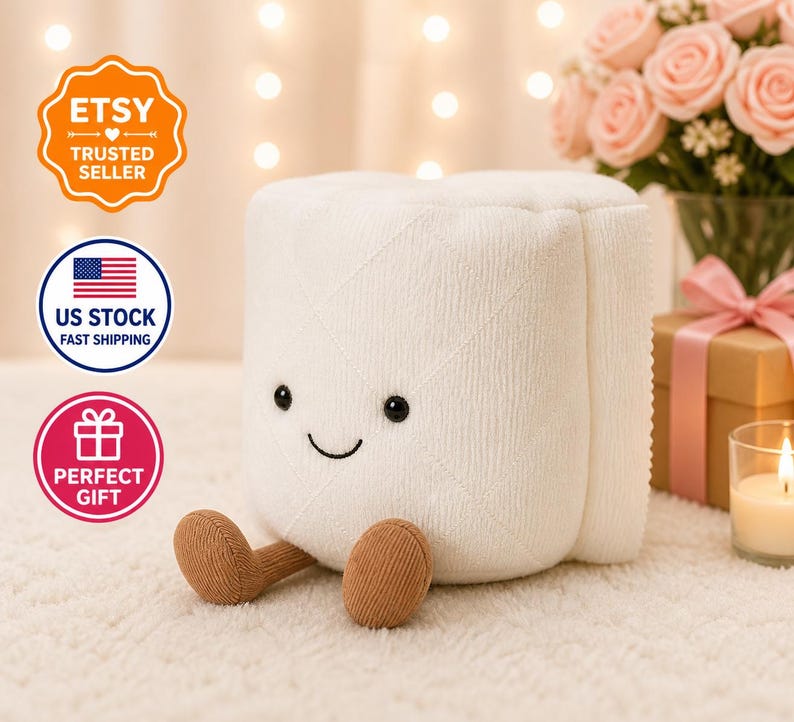 May include: A white plush toy shaped like a roll of toilet paper with a smiling face and brown feet. The toy has black eyes and a stitched smile. The image also includes a bouquet of pink roses and a gift box.