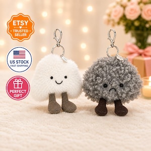 May include: Two plush cloud-shaped keychains. One is white with a smiling face, the other is gray with a sad face. Both have brown feet and silver key rings. The keychains are designed to look like cute, fluffy clouds.