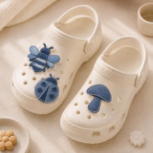 May include: A pair of off-white clogs with decorative denim patches. One clog features a bee and ladybug, the other a mushroom. The clogs have multiple ventilation holes and a pivoting heel strap.