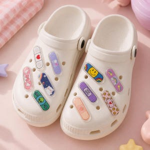 May include: A pair of white clogs adorned with colorful, decorative bandage-shaped charms. The charms feature various designs, including a smiley face, a rainbow, and an alien. The clogs have a classic design with ventilation holes.