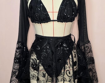 May include: Black lingerie set featuring a halter-neck bra, long-sleeved sheer top, and a wrap skirt. The bra and skirt are embellished with sequins and floral lace. The sheer top has bell sleeves. The set is displayed on a mannequin.