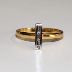 Puede incluir: Un anillo de oro con una barra vertical plateada con tres peque&ntilde;os diamantes rectangulares. La banda del anillo es de color oro pulido, y los diamantes est&aacute;n engastados en metal plateado.