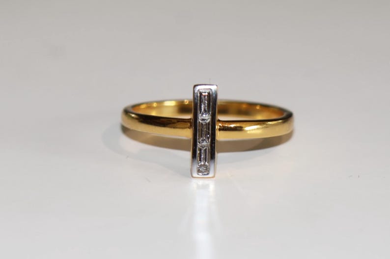 Puede incluir: Un anillo de oro con una barra vertical plateada con tres peque&ntilde;os diamantes rectangulares. La banda del anillo es de color oro pulido, y los diamantes est&aacute;n engastados en metal plateado.