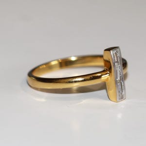 Puede incluir: Un anillo de oro con un acento rectangular con diamantes. La banda del anillo es de oro pulido, y el acento rectangular presenta una fila de peque&ntilde;os diamantes transparentes. El anillo est&aacute; sobre un fondo blanco.