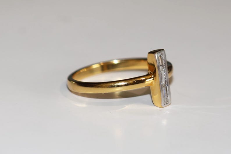 Puede incluir: Un anillo de oro con un acento rectangular con diamantes. La banda del anillo es de oro pulido, y el acento rectangular presenta una fila de peque&ntilde;os diamantes transparentes. El anillo est&aacute; sobre un fondo blanco.