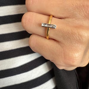 Puede incluir: Un anillo de oro con un centro rectangular de diamantes de talla baguette. El anillo se lleva en un dedo, sobre un fondo de tela a rayas blancas y negras. La banda del anillo es de oro pulido.