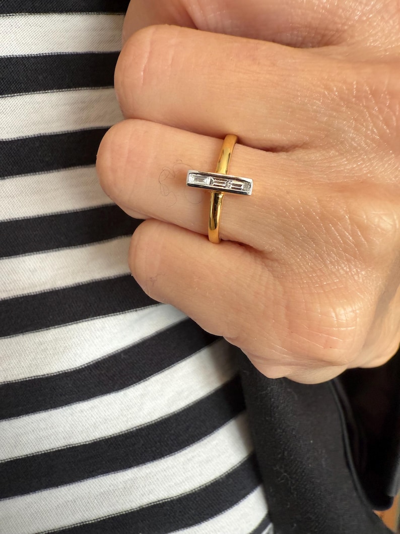 Puede incluir: Un anillo de oro con un centro rectangular de diamantes de talla baguette. El anillo se lleva en un dedo, sobre un fondo de tela a rayas blancas y negras. La banda del anillo es de oro pulido.