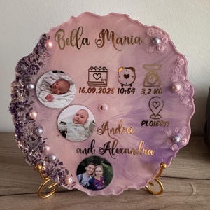 Op de afbeelding: Decoratieve harsplaat in roze en paars met de namen "Bella Maria" en "Andrei and Alexandra" in goud. Inclusief babyfoto's, geboortedetails en decoratieve elementen. Ondersteund door een gouden standaard.