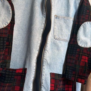 Peut inclure: Une veste en jean bleu clair avec une fermeture éclair frontale, un col et des manches longues. La veste présente des empiècements en tissu à carreaux rouges, verts et noirs sur les manches et les poches avant, ainsi que des coudières en jean.
