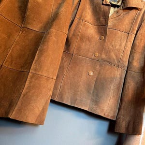 May include: A tan suede two-piece set consisting of a long-sleeved button-front jacket and a matching skirt. Both pieces feature a visible grid-like panel construction with stitched seams throughout the garment. The jacket has a collar and buttons.