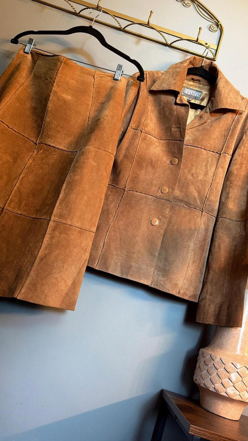 May include: A tan suede two-piece set consisting of a long-sleeved button-front jacket and a matching skirt. Both pieces feature a visible grid-like panel construction with stitched seams throughout the garment. The jacket has a collar and buttons.