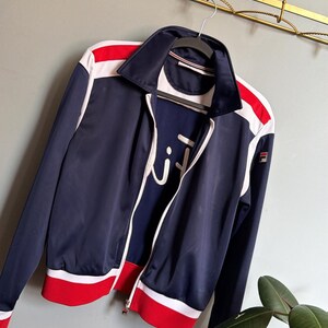 Op de afbeelding: Marineblau sportjack met witte en rode accenten. De jas heeft een volledige ritssluiting, een witte kraag en het merklogo. Het hangt aan een hanger.