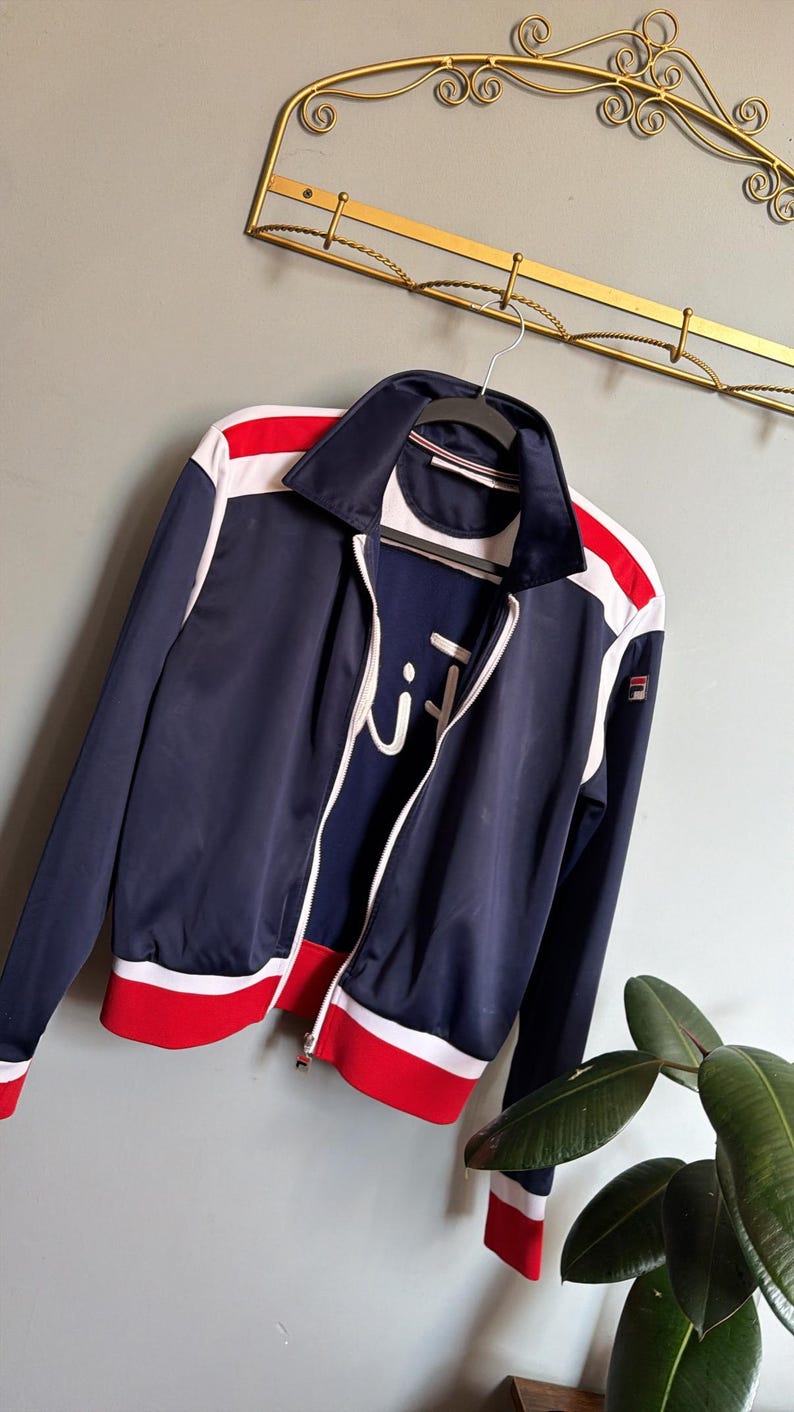 Op de afbeelding: Marineblau sportjack met witte en rode accenten. De jas heeft een volledige ritssluiting, een witte kraag en het merklogo. Het hangt aan een hanger.