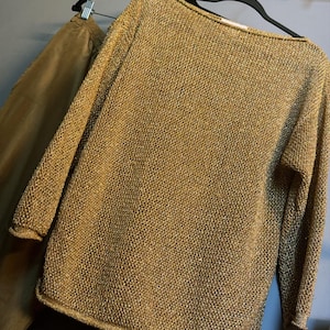May include: A gold-coloured knitted sweater with long sleeves and a boat neck is displayed on a black hanger. The sweater has a textured appearance and is paired with brown trousers. A decorative gold-coloured wall-mounted rack is visible.