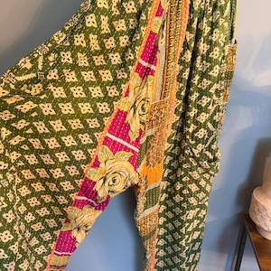 May include: Loose-fitting pants featuring a patchwork design with olive green fabric patterned with small white geometric shapes, accented by vertical panels of bright pink and yellow floral prints, finished with visible white running stitch details.
