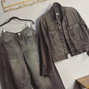 May include: A matching denim jacket and high-waisted jeans set in a dark gray wash. The jacket has button closures and pockets. The jeans have a relaxed fit. The clothing is hanging on a wall with a decorative gold metal rack.