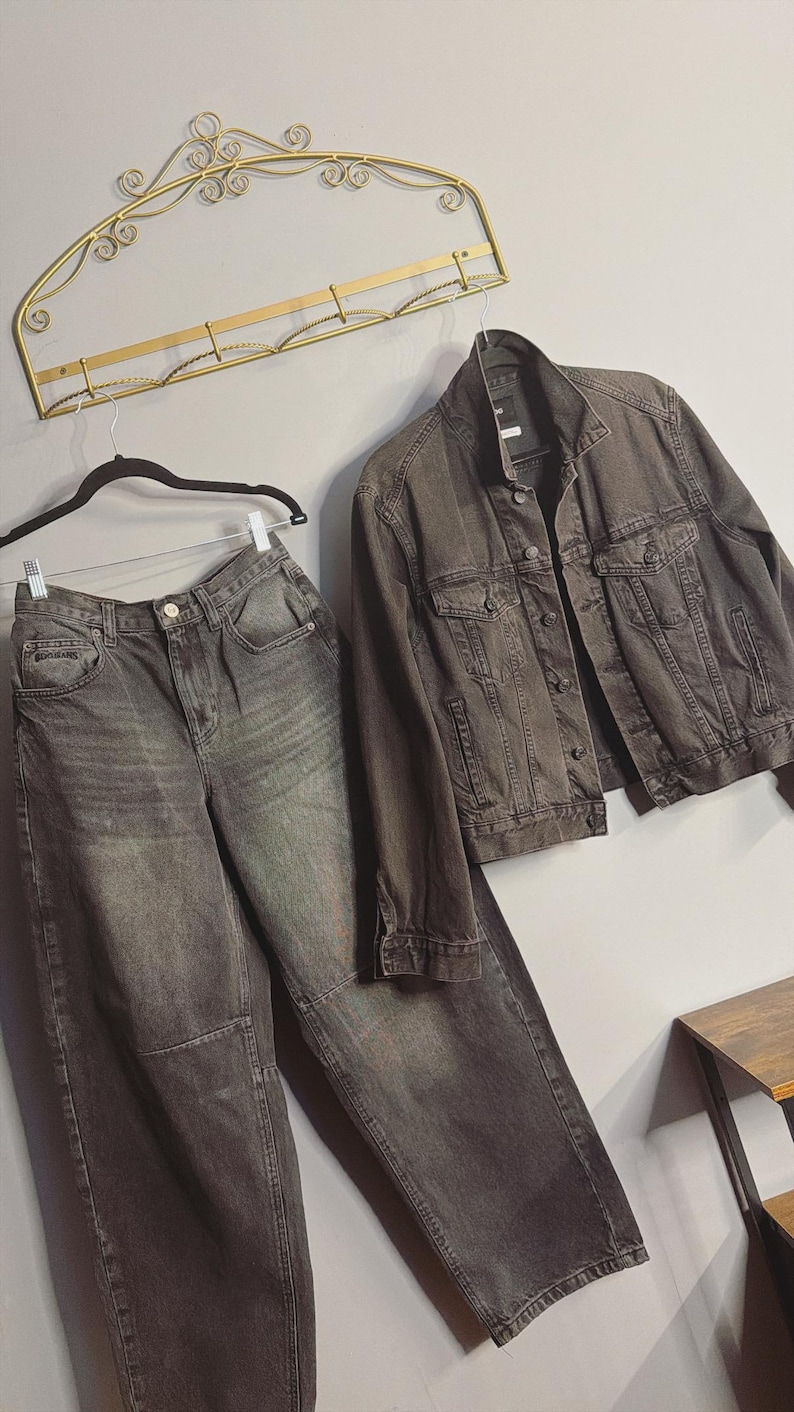 May include: A matching denim jacket and high-waisted jeans set in a dark gray wash. The jacket has button closures and pockets. The jeans have a relaxed fit. The clothing is hanging on a wall with a decorative gold metal rack.