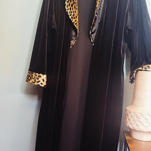May include: A long, black velvet robe featuring a wide shawl collar and sleeve cuffs trimmed with a brown and black leopard print pattern. The robe is open at the front, revealing a solid black, floor-length garment worn underneath.