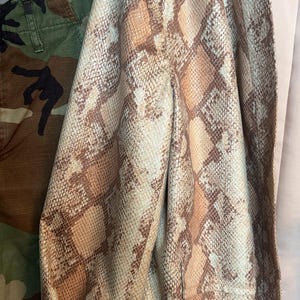 May include: A snakeskin-patterned blazer in shades of tan, brown, and cream hangs on a black hanger. The blazer has a light pink lining and rolled-up sleeves. Camouflage pants are visible on the left side of the image.