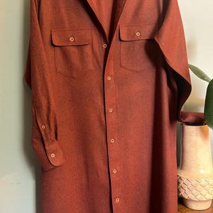 May include: A long-sleeved, button-down shirt in a solid, deep rust-orange color. The shirt features a classic collar, two chest pockets with buttoned flaps, and a full-length front button closure. The fabric has a subtle, fine-grained texture.