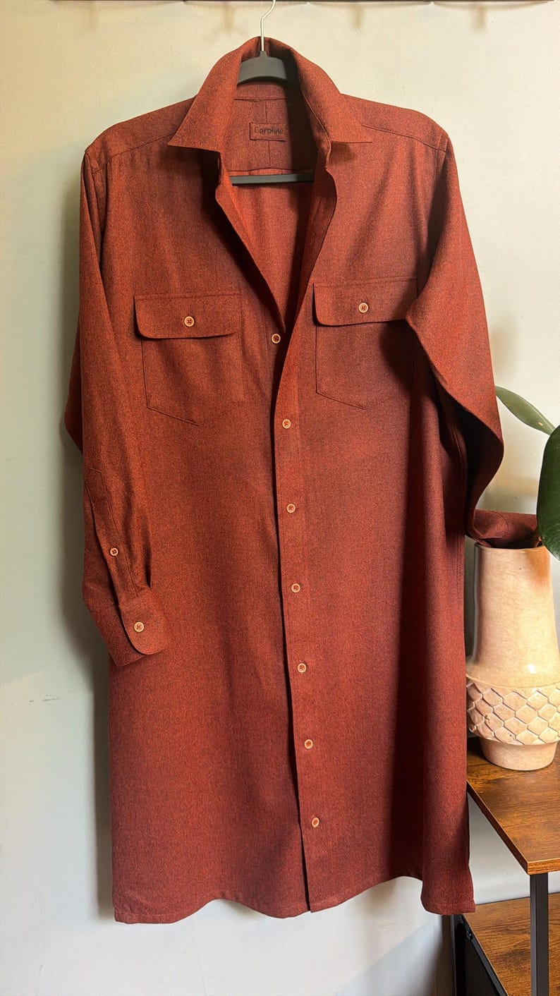 May include: A long-sleeved, button-down shirt in a solid, deep rust-orange color. The shirt features a classic collar, two chest pockets with buttoned flaps, and a full-length front button closure. The fabric has a subtle, fine-grained texture.