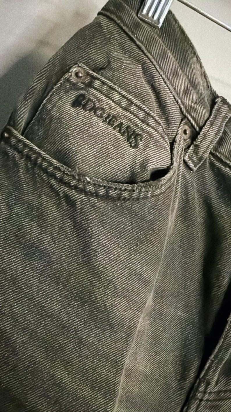 May include: Close-up of a pair of faded olive green jeans. The jeans feature a classic five-pocket design with the brand name "BDG JEANS" embroidered on the back pocket. The fabric has a textured appearance.