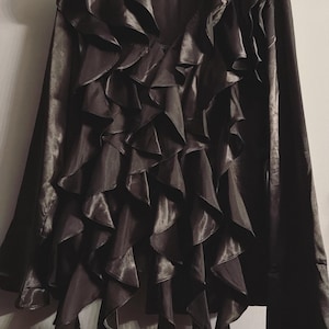 May include: A dark gray blouse with a V-neck and long sleeves. The front of the blouse is adorned with multiple layers of ruffled fabric. The blouse is hanging on a black hanger against a light gray wall.