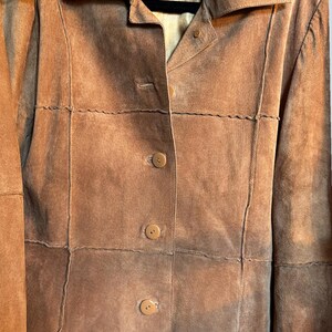 May include: A tan suede jacket featuring a pointed collar, long sleeves, and a front button closure with three visible buttons. The garment includes horizontal and vertical panel stitching details and a label reading ORIGINAUX PABLO MONTREAL CANADA.