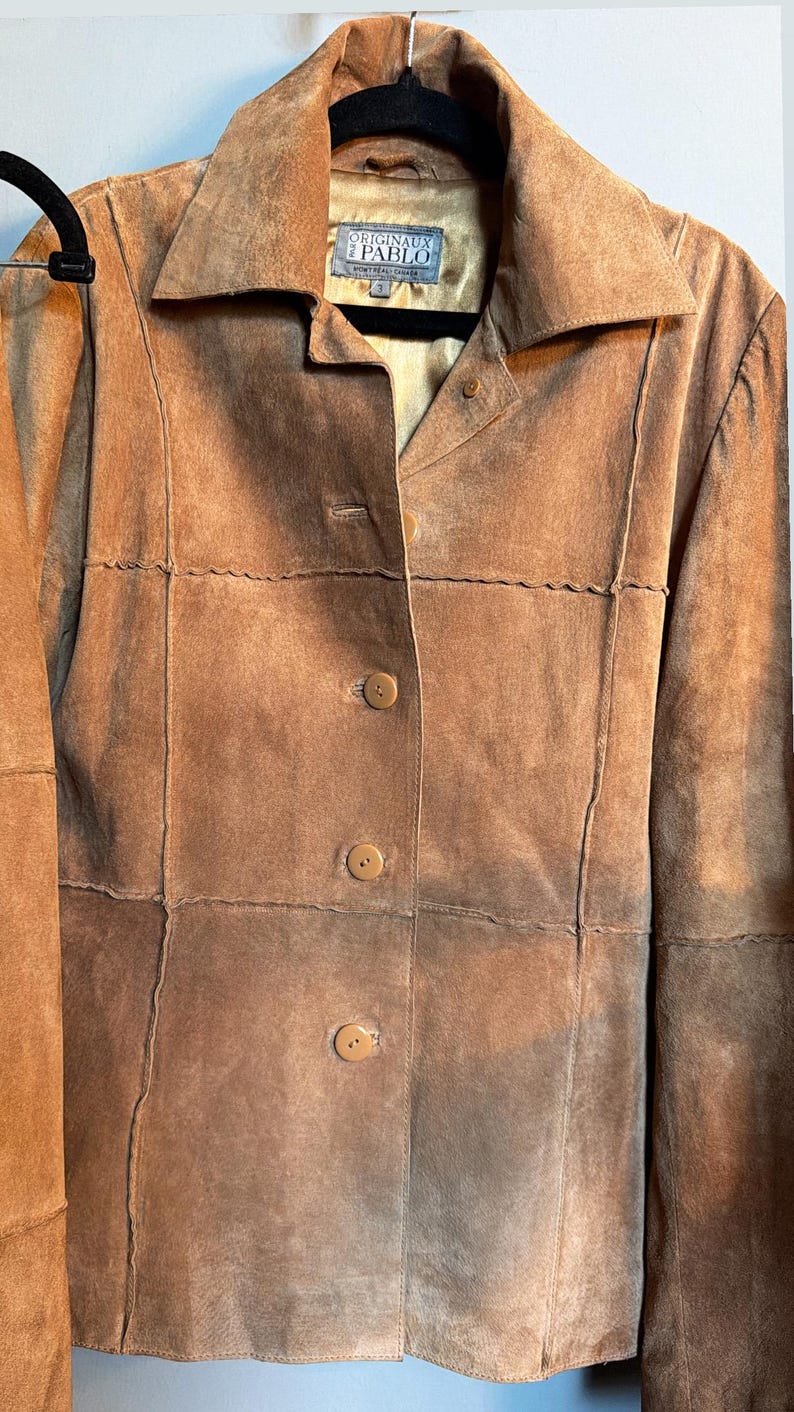 May include: A tan suede jacket featuring a pointed collar, long sleeves, and a front button closure with three visible buttons. The garment includes horizontal and vertical panel stitching details and a label reading ORIGINAUX PABLO MONTREAL CANADA.