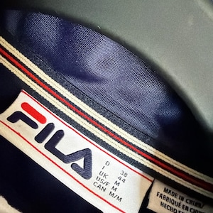 Op de afbeelding: Close-up van de kraag van een marineblauwe FILA-jas. De kraag heeft een wit, rood en marineblauw streepontwerp. Het FILA-logo staat op een label, met maatdetails.