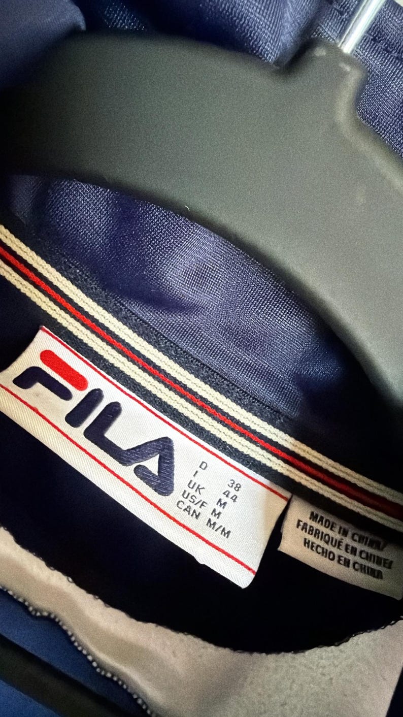 Op de afbeelding: Close-up van de kraag van een marineblauwe FILA-jas. De kraag heeft een wit, rood en marineblauw streepontwerp. Het FILA-logo staat op een label, met maatdetails.