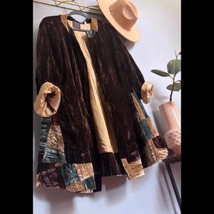May include: A brown velvet coat with patchwork details in shades of green, tan, and brown. The coat has a tan lining and rolled-up sleeves. A tan hat with a beaded band rests on a gold-colored coat rack.