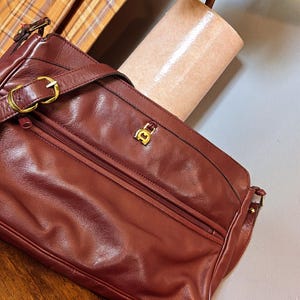 May include: A burgundy leather shoulder bag with a gold-tone buckle and zipper. The bag has a gold logo on the front and a matching burgundy belt with a gold buckle. A plaid fabric is visible in the background.