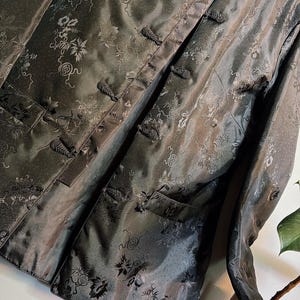 Peut inclure: Veste à manches longues gris foncé avec un col mao et des fermetures à brandebourgs. La veste présente un motif floral et deux poches avant. Le tissu a un éclat subtil, et la veste semble être en soie ou dans une matière similaire.