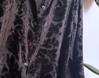 May include: A black long-sleeve button-down shirt featuring a tonal velvet floral pattern throughout. The garment has a pointed collar, a front button placket with dark red buttons, and a semi-sheer base fabric with a textured, raised floral design.