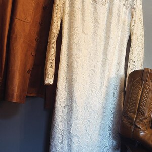 May include: A long-sleeved white lace dress with a round neckline, a tan leather jacket with buttons, and a brown leather cowboy boot with decorative stitching. The dress features a floral lace pattern throughout and hangs on a black hanger.