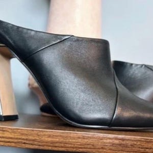 May include: Black leather mules with a pointed toe and a gold-colored, geometric heel. The shoes are displayed on a wooden shelf, showcasing their sleek design and modern aesthetic.