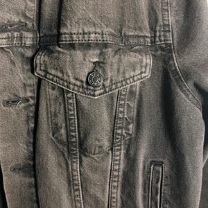 May include: Close-up of a charcoal-colored denim jacket. The jacket features a button-down front, a chest pocket with a button closure, and a small side pocket. The fabric has a textured appearance.