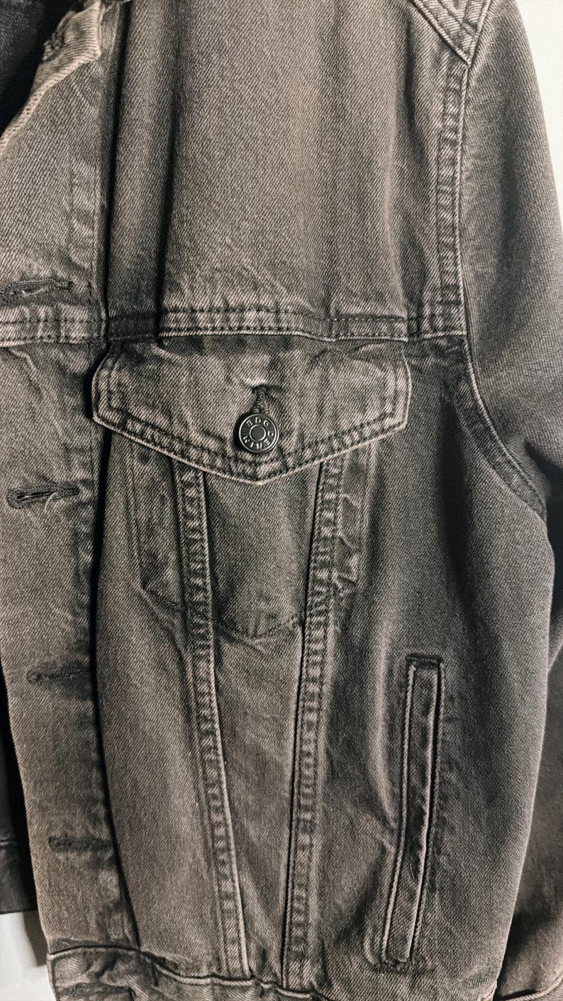 May include: Close-up of a charcoal-colored denim jacket. The jacket features a button-down front, a chest pocket with a button closure, and a small side pocket. The fabric has a textured appearance.