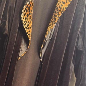 May include: A long, dark brown velvet robe featuring a wide shawl collar and sleeve cuffs lined with a bold, black and orange leopard print pattern. The garment is open at the front, showcasing the smooth, dark brown texture of the velvet fabric.