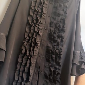 May include: A long-sleeved black button-down shirt featuring a prominent vertical ruffled panel down the center front. The shirt includes a classic collar and tiered ruffled cuffs on the sleeves, constructed from a lightweight, semi-sheer fabric.