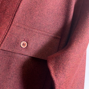 May include: A long-sleeved shirt in a deep burgundy red color with a textured, heathered fabric finish. The shirt features a front chest pocket with a single, centered, light-colored button that has four small holes in the middle of the button.